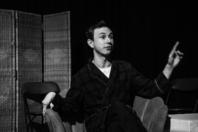 Black & white image of Oliver as GOD. He is sitting in a shell chair mid gesture as he addresses the audience.